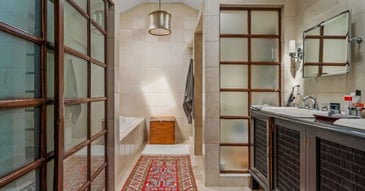 Bathroom with beige tile, wood-framed glass partitions, a soaking tub, and dark wood vanity with dual sinks.
